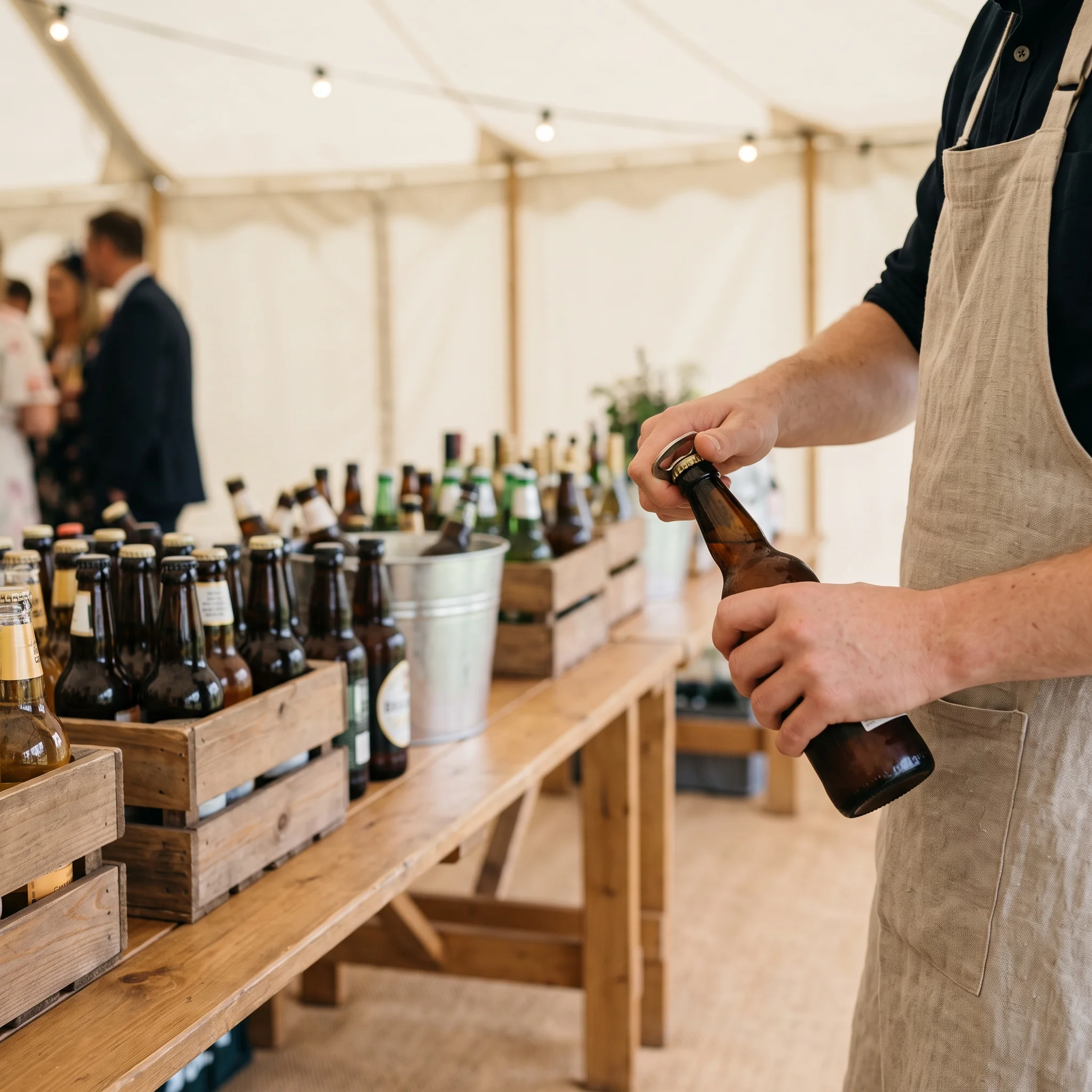 A trestle bar in a marquee with rows of beer and cider bottles and a server's hands opening one