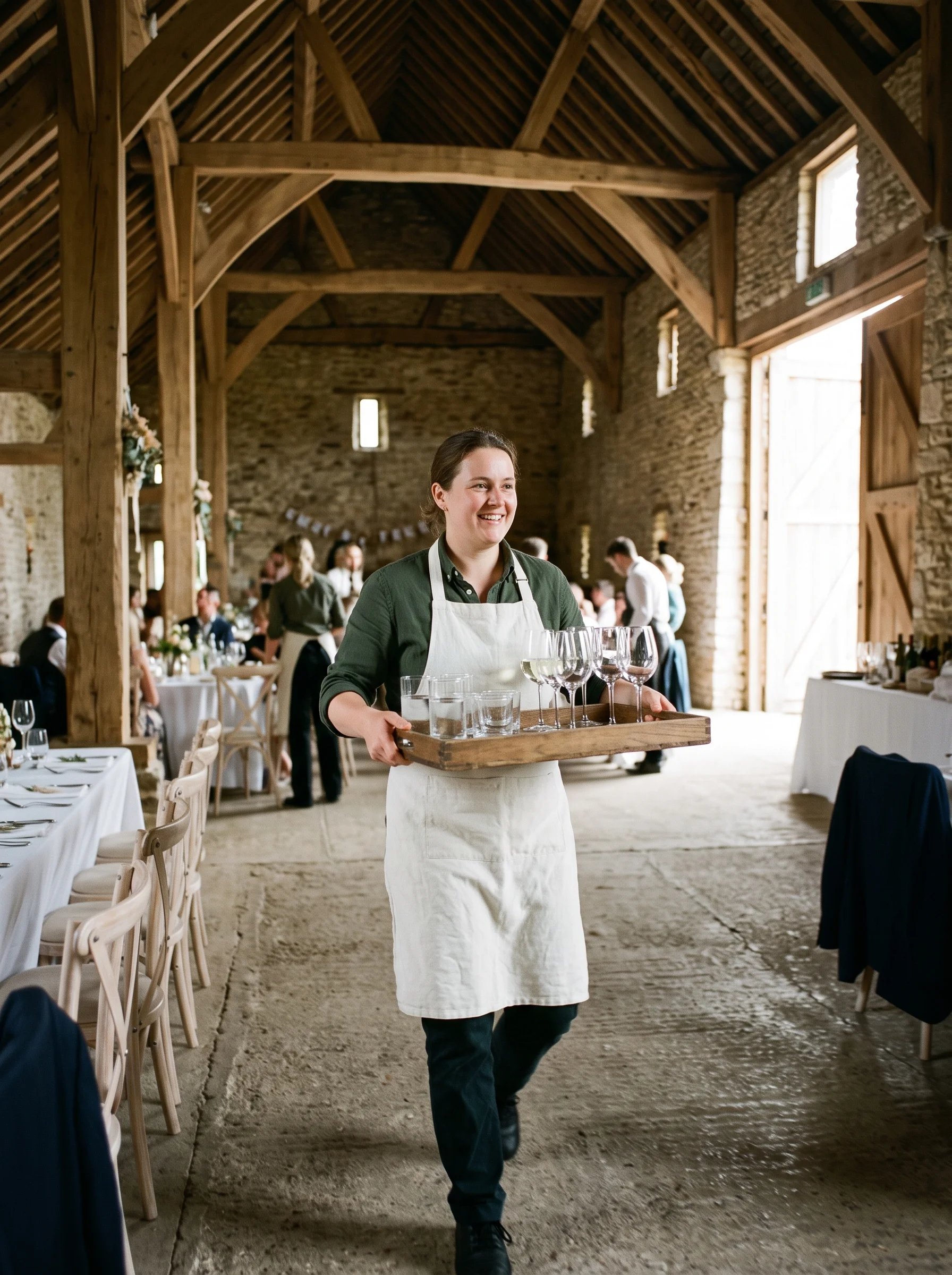 A server in an apron carrying a tray of glasses across a daylit barn with guests blurred around him