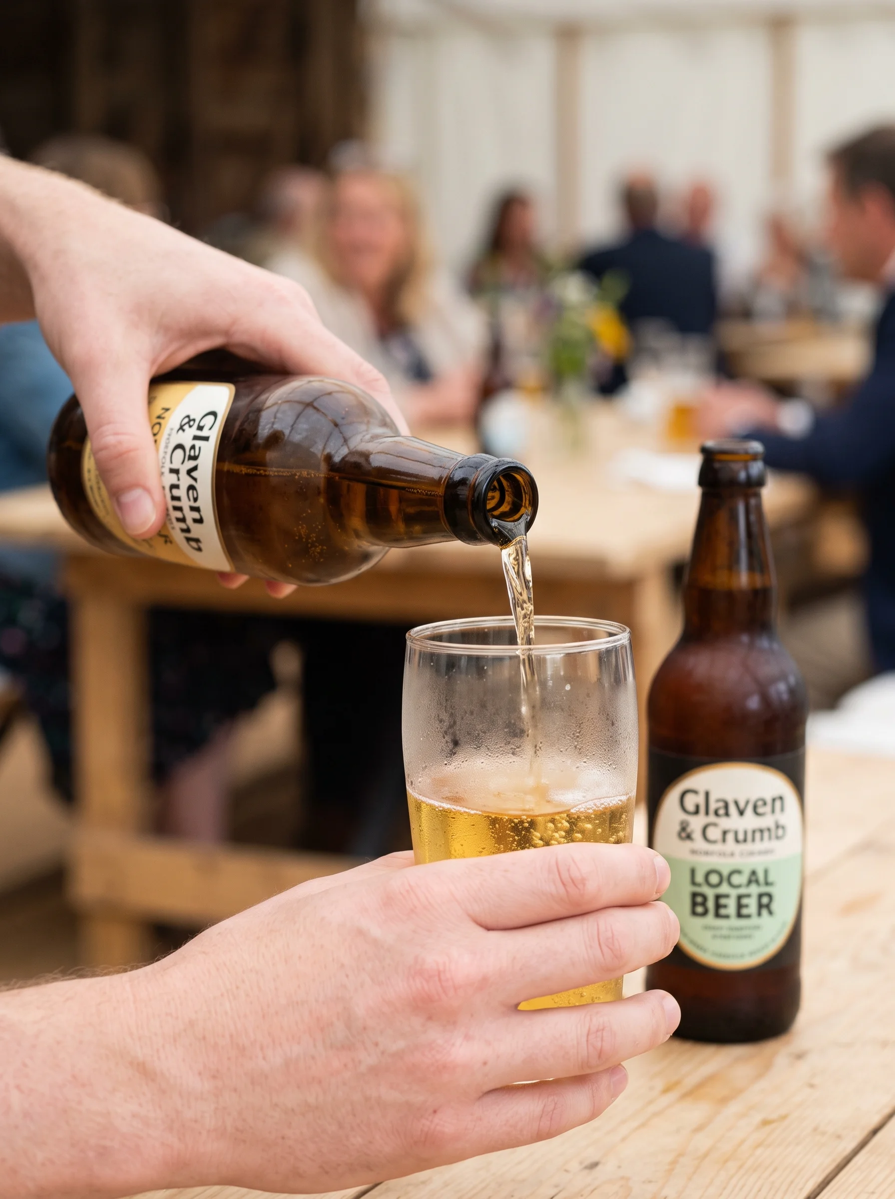 A hand pouring a beverage from a brown bottle into a dewed glass on a pale wooden table, with another bottle behind and people blurred in the background.